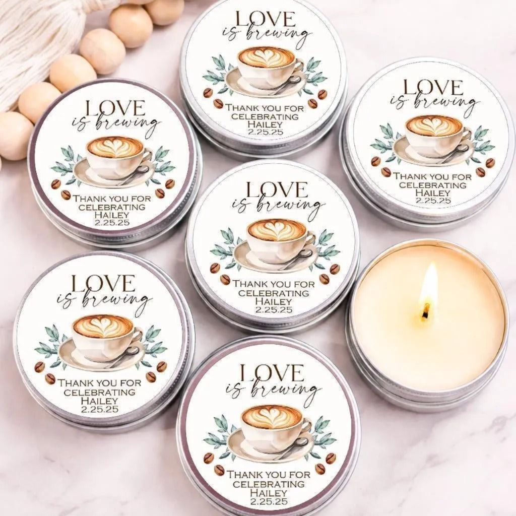 a collection of six silver tin candles with white labels, each featuring a coffee cup and the text "LOVE is brewing" along with a heart symbol. The candles are arranged on a marble surface, with some of them lit, creating a warm and cozy atmosphere.