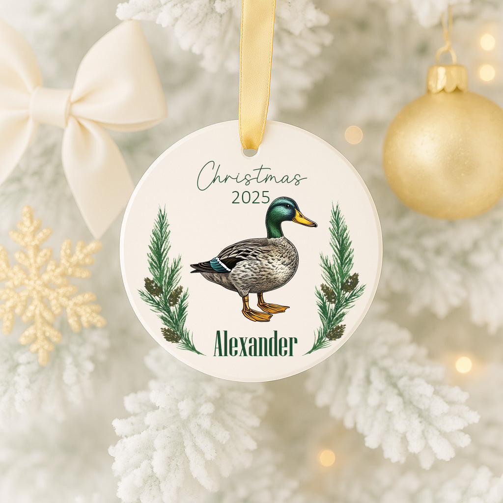 A white Christmas ornament with a duck image and the name "Alexander" is hanging on a tree.