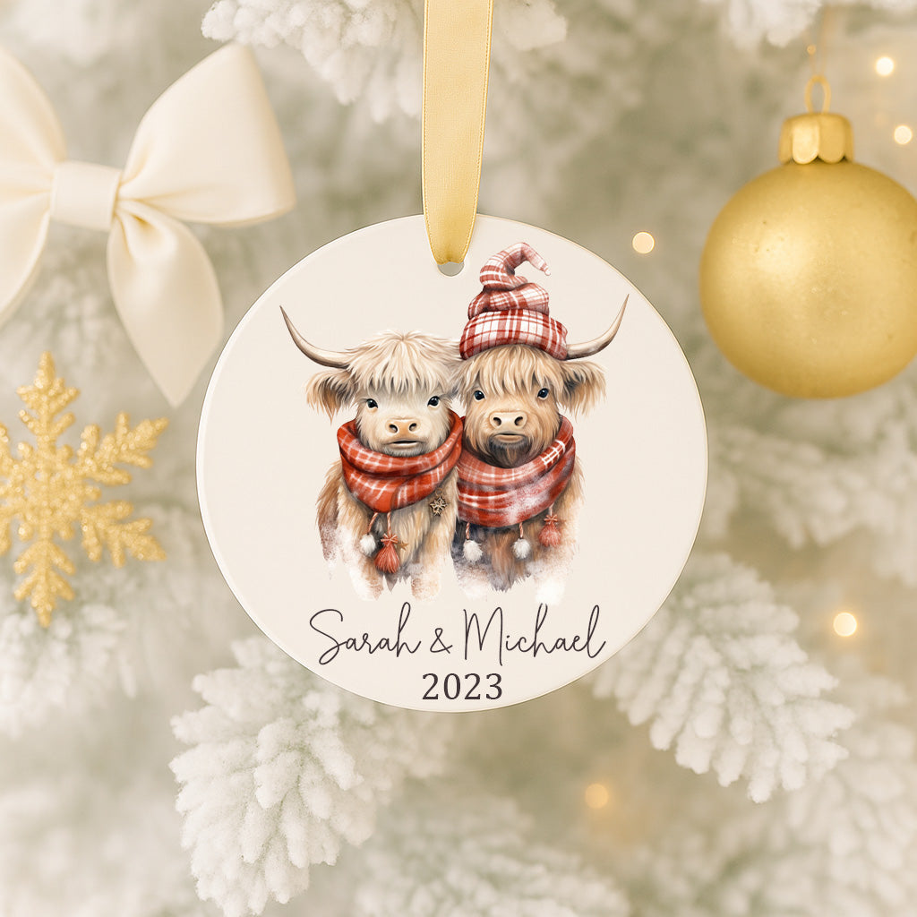A white ornament with two cows wearing red and white plaid hats, and the text "Sarah & Michael" and "2023" printed on it, hanging on a Christmas tree.