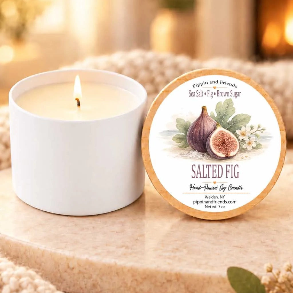 a white candle with a label that reads "SALTED FIG" and "Pippin and Friends" next to a white candle holder.