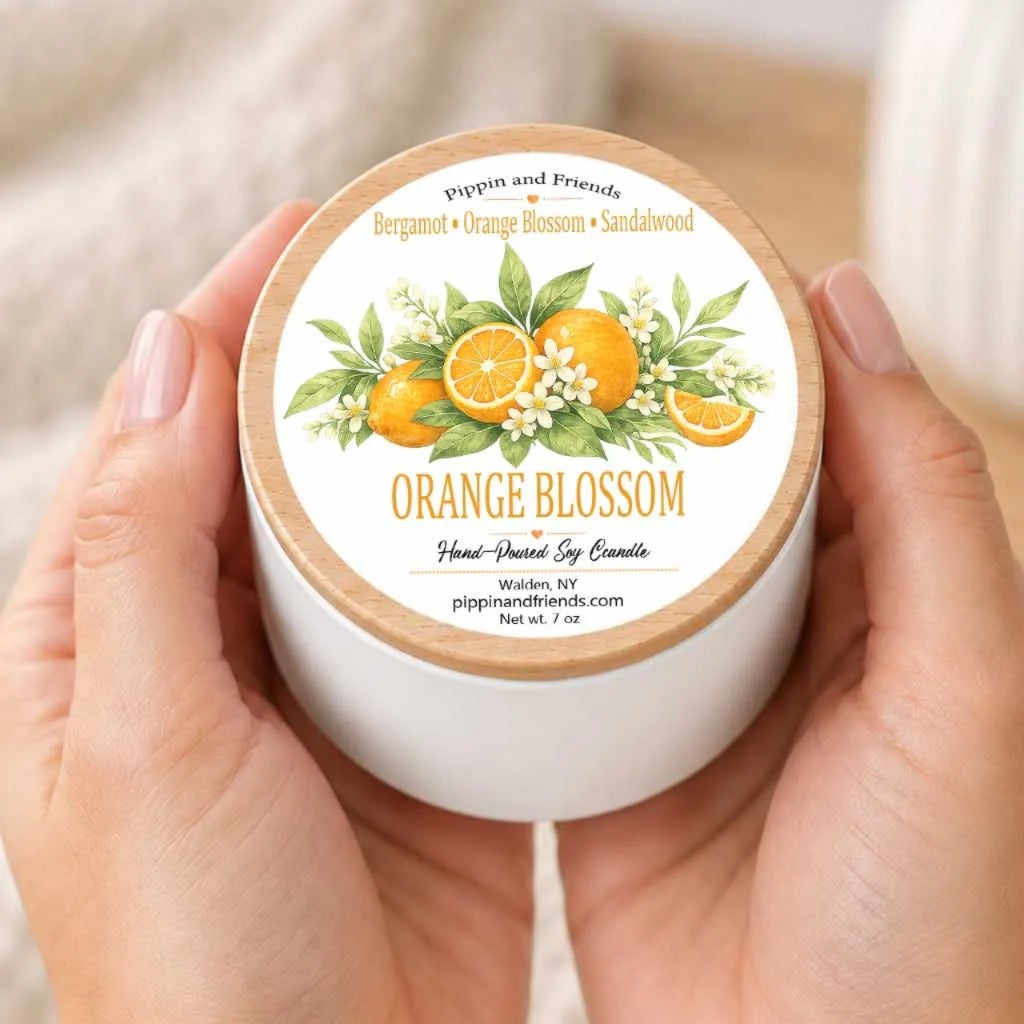 A person's hands holding a white container with a label that features an illustration of oranges and flowers, along with the text "Orange Blossom" and "Pippin and Friends Bergamot Orange Blossom Sandalwood".