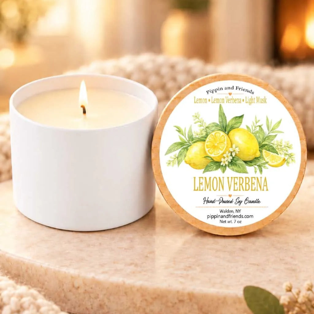 a white candle with a label that reads "Lemon Verbena" and "Pippin and Friends" on it, placed on a wooden surface next to a white candle holder.