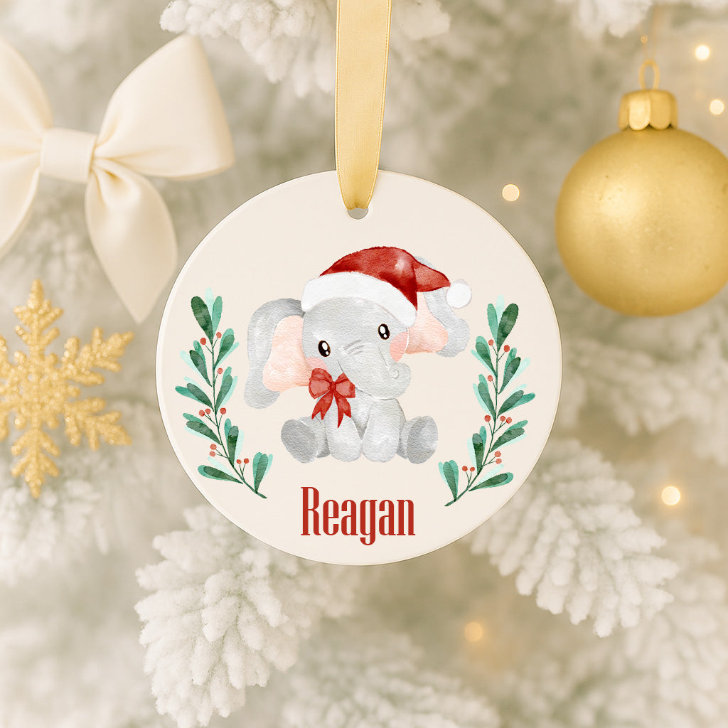 A white ornament with a red bow and a small elephant wearing a Santa hat, surrounded by green leaves and berries, hangs on a Christmas tree.