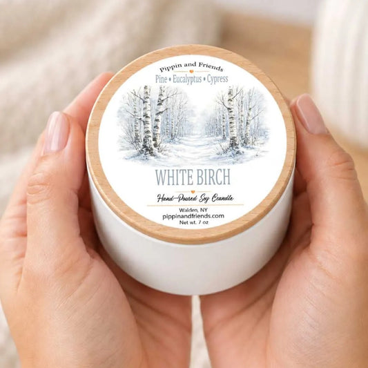 A person's hands holding a white candle container with a label depicting a snowy forest scene.