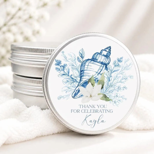 A round tin lid with a blue and white seashell design and white flowers, placed on a white cloth.