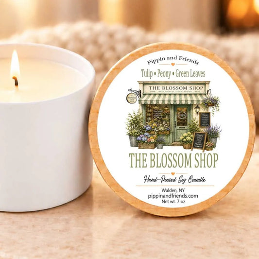 a white candle with a label that reads "The Blossom Shop" and "Hand-Poured Soy Candle" on a table.