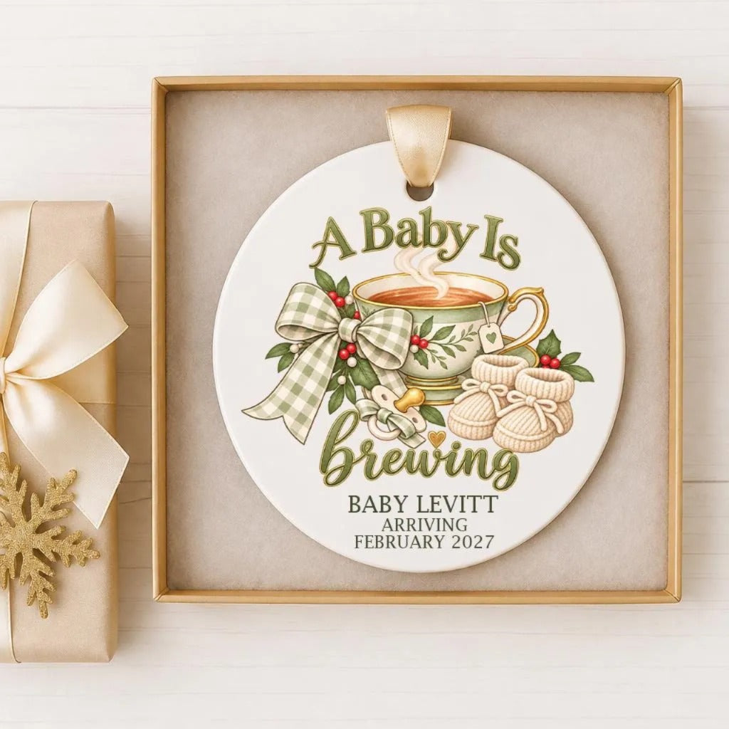 A decorative ornament with the text "A Baby Is Brewing" and illustrations of a cup of tea, baby booties, and a ribbon.