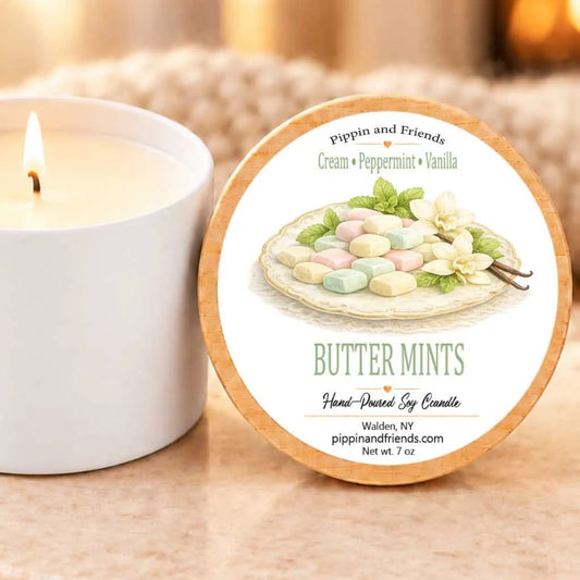 a white candle with a label that reads "Butter Mints" and "Hand-poured by Candle", placed next to a white candle holder.