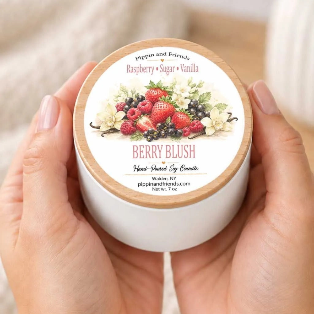 A person's hands holding a white container with a label featuring a floral design and the text "Berry Blush" and "Pippin and Friends Raspberry + Sugar + Vanilla".
