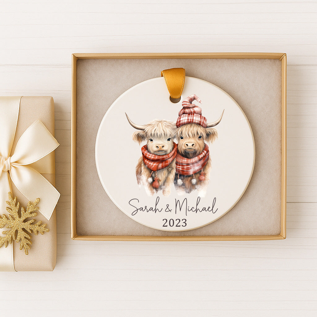 A ceramic ornament featuring two cows wearing winter clothing, with the text "Sarah & Michael" and the year "2023" printed on it. The ornament is displayed in a gold box, and there is a gift box with a gold ribbon next to it.