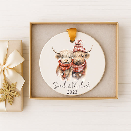 A ceramic ornament featuring two cows wearing winter clothing, with the text "Sarah & Michael" and the year "2023" printed on it. The ornament is displayed in a gold box, and there is a gift box with a gold ribbon next to it.