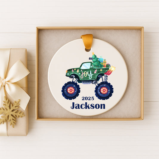 A white ornament with a green monster truck and the word "Joy" on it, placed inside a gold box.