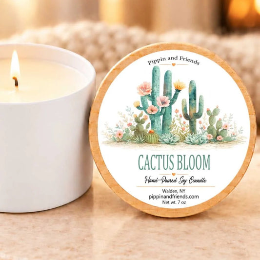 a white candle with a cactus design and a round label that reads "Cactus Bloom Hand-Dipped Soy Candle Walden, NY pippinandsrands.com 7 oz" placed next to it.