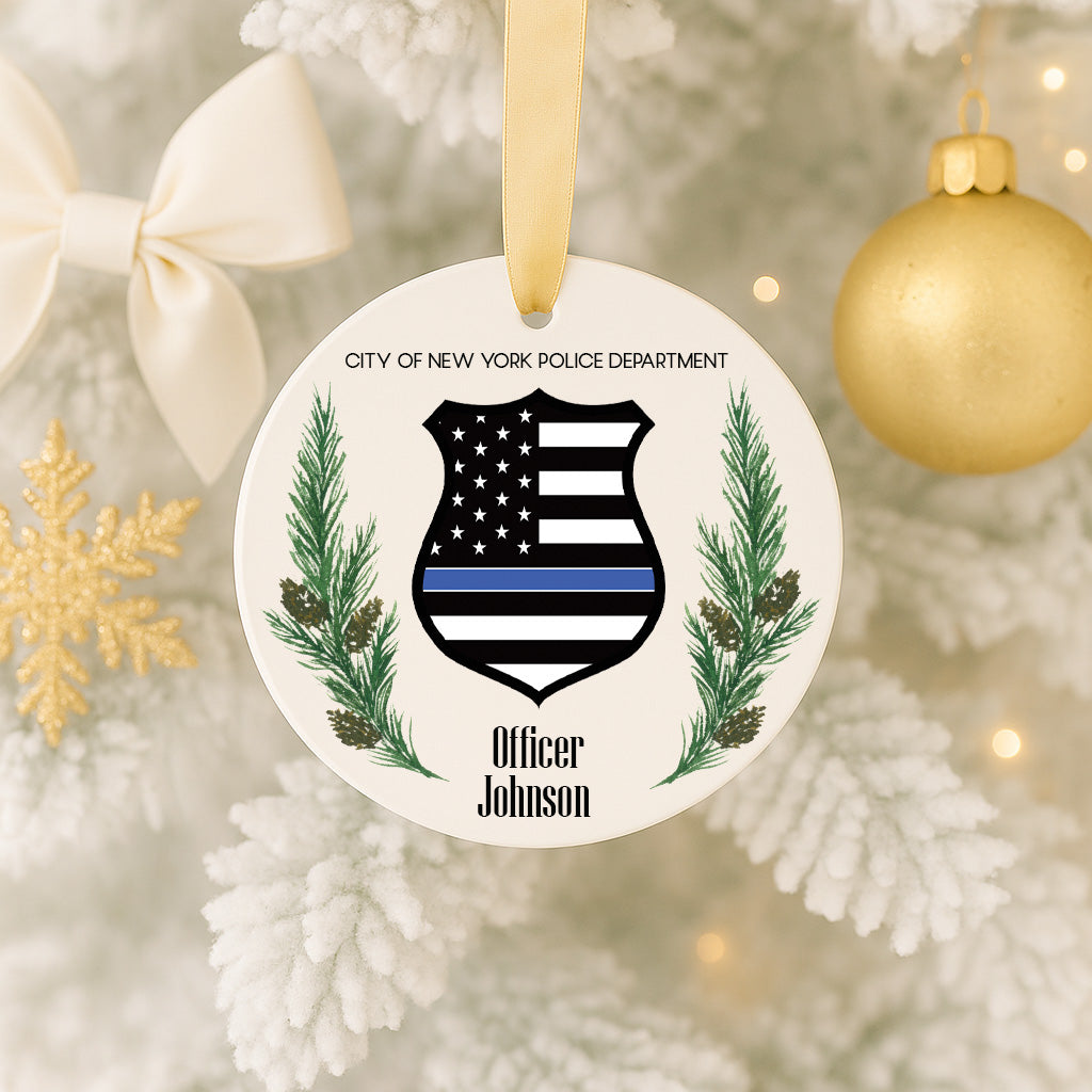 A white ornament with a black and white shield design, featuring a thin blue line, pine branches, and pinecones, hangs on a Christmas tree.