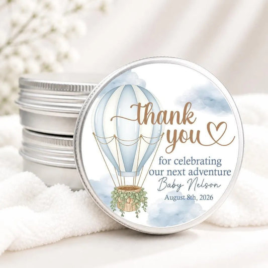 A round tin lid with a decorative design featuring a hot air balloon and the text "Thank you for celebrating our next adventure Baby Nelson August 8th, 2026".