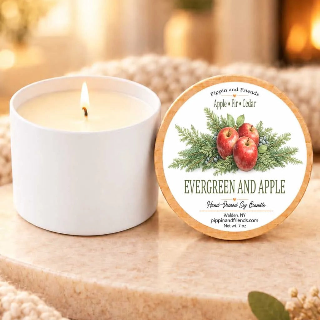 a white candle with a label that reads "EVERGREEN AND APPLE" and "Pippin and Friends Apple + Cedar" on a wooden surface. The candle is accompanied by a small white ceramic container with a similar label.