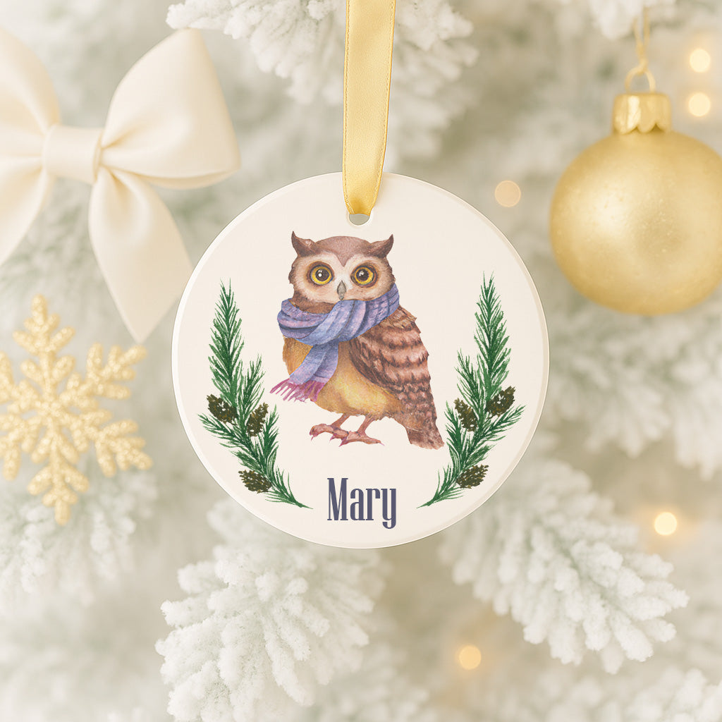 A white ornament with a picture of an owl wearing a scarf and surrounded by pine branches, with the name "Mary" written below the image.