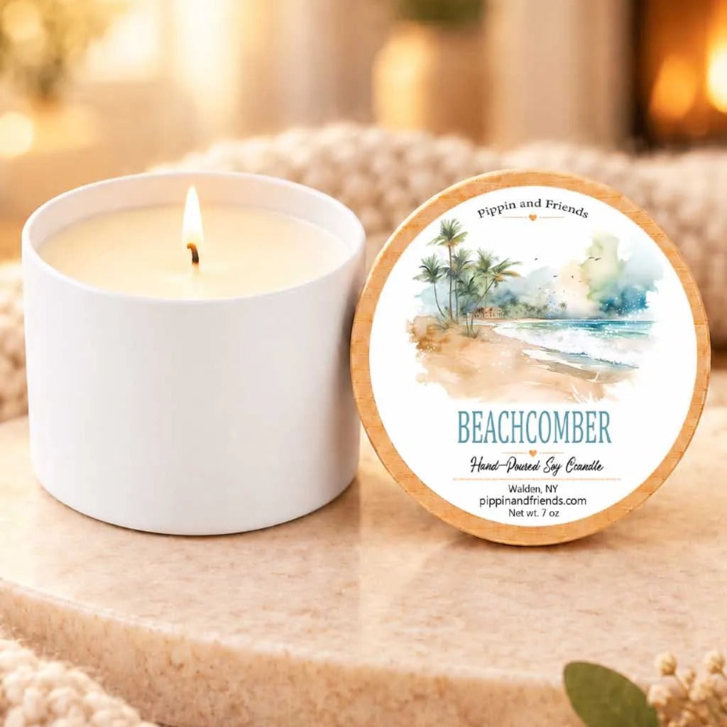 a white candle labeled "Beachcomber" placed on a wooden surface, with a blurred background that includes a fireplace and a window.