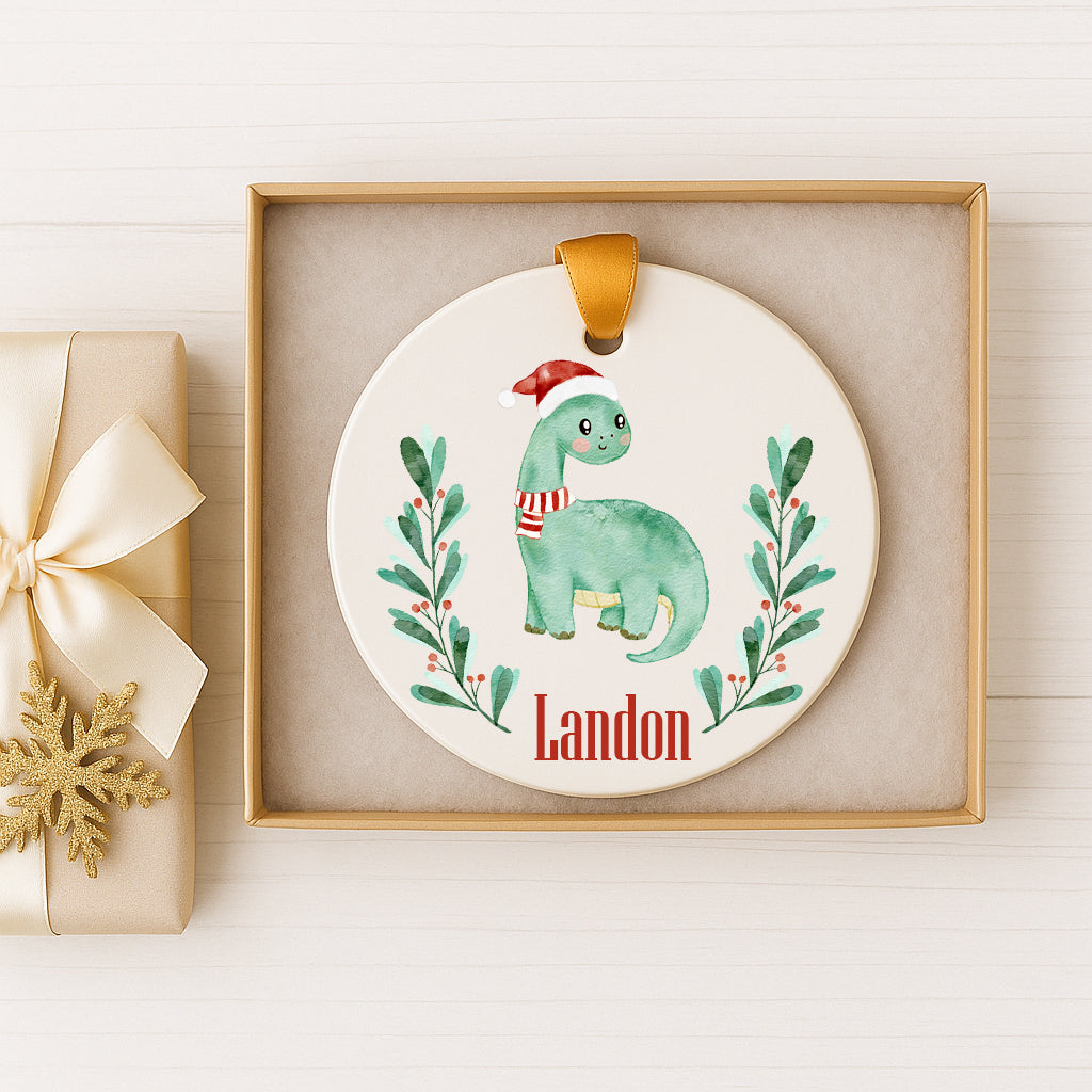A ceramic ornament featuring a green dinosaur wearing a Santa hat, surrounded by green leaves and berries, with the name "Landon" written on it. The ornament is displayed in a wooden frame, and there is a gift box with a gold bow next to it.