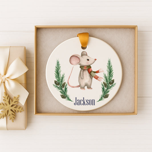 A white ceramic ornament with a mouse and pine tree design, featuring the name "Jackson" written on it, is displayed in a gold box.