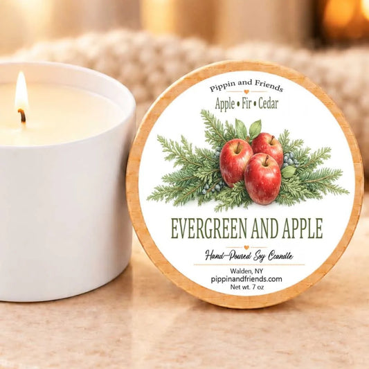 a white candle with a label that reads "EVERGREEN AND APPLE Hand-Drawn Soy Candle Walden, NY pippinandfriends.com net wt 7 oz". The candle is placed next to a white candle holder, creating a cozy and inviting scene.