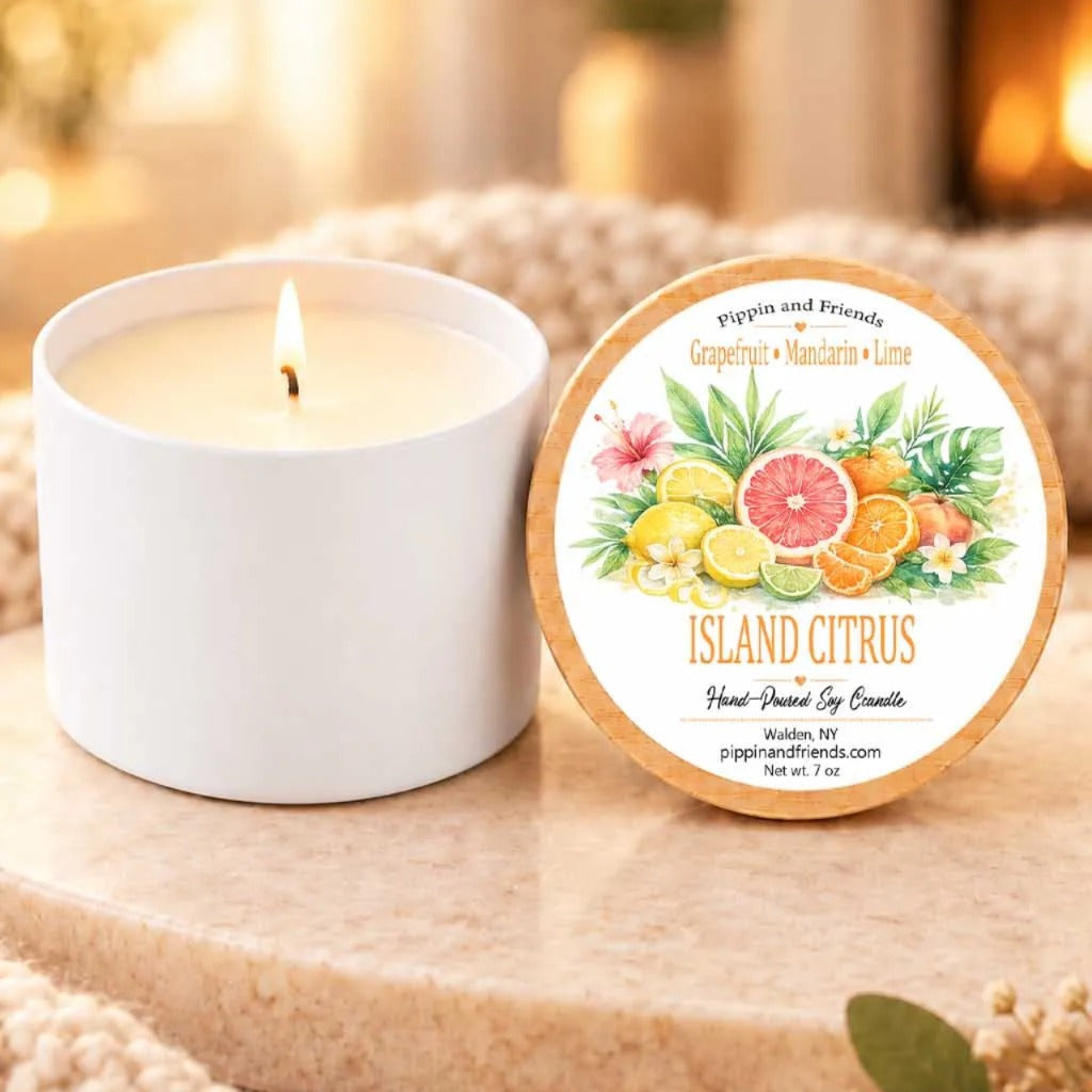 a white candle with a label that reads "Island Citrus Pippin and Friends Grapefruit & Mandarin Lime" and "Hand Picked by Friends" on a wooden surface. Next to the candle is a white ceramic container with a lid, and there are some greenery and a small plant in the background.