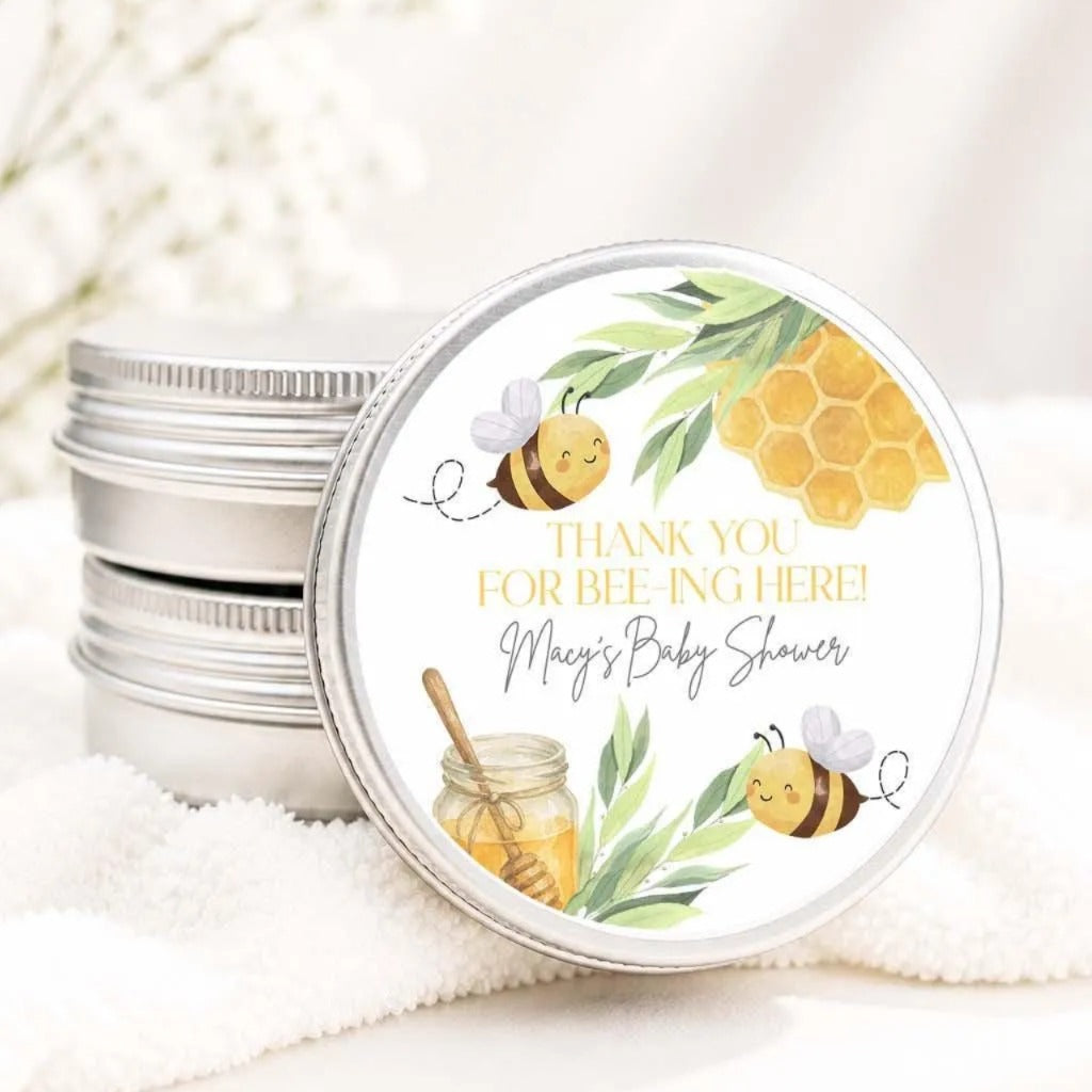 A round tin lid with a label featuring illustrations of bees and honey, along with the text "Thank you for beeing here! Macy's Baby shower".