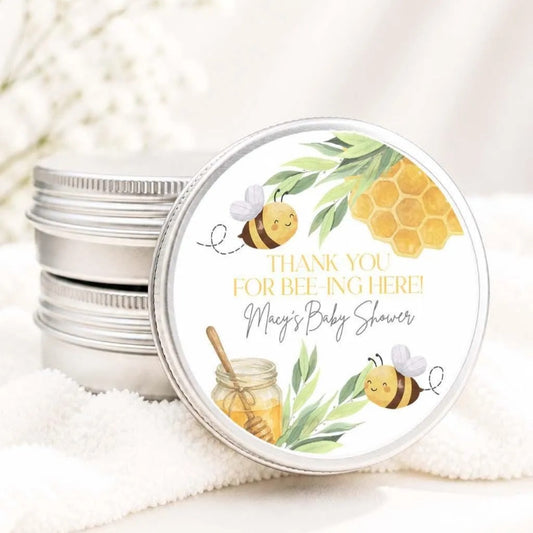 A round tin lid with a label featuring illustrations of bees and honey, along with the text "Thank you for beeing here! Macy's Baby shower".