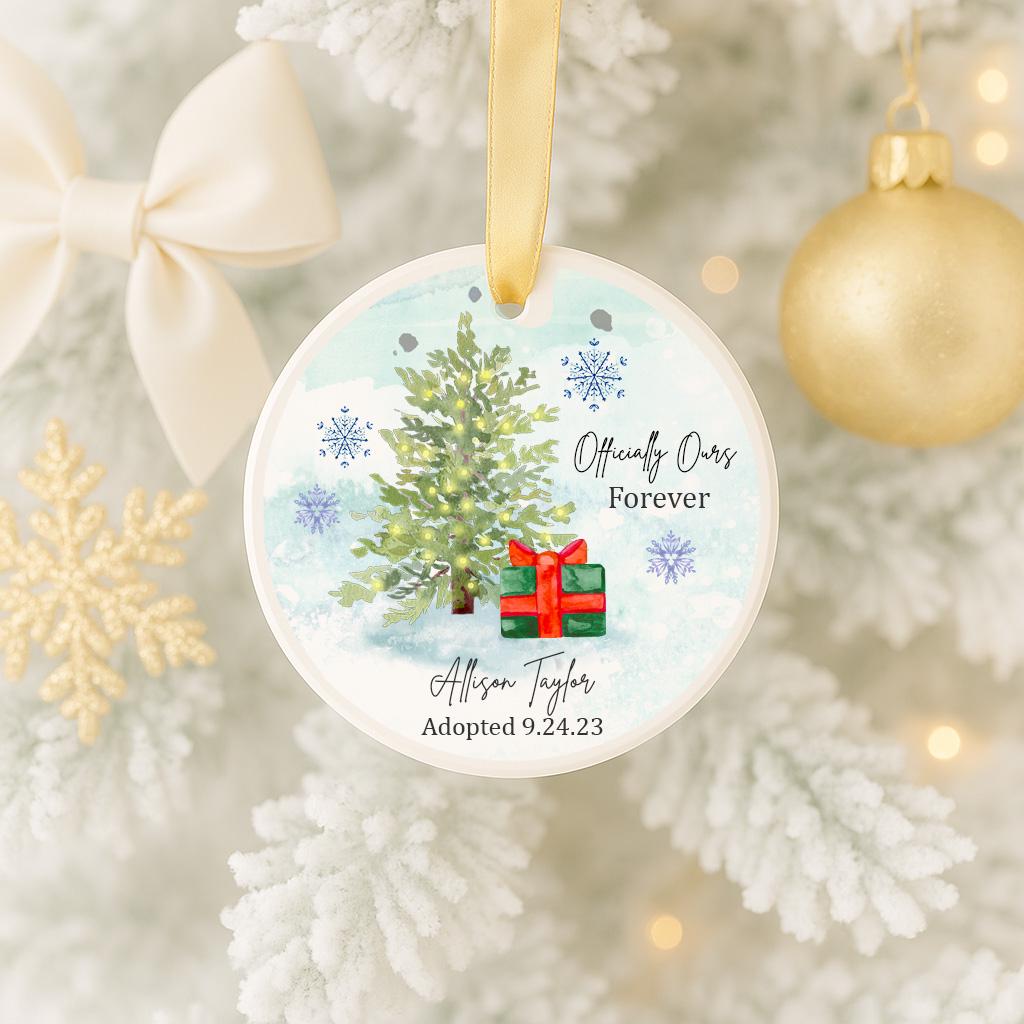 A white Christmas tree ornament with a red bow and a green gift box, hanging on a tree with a gold ornament and snowflakes in the background.