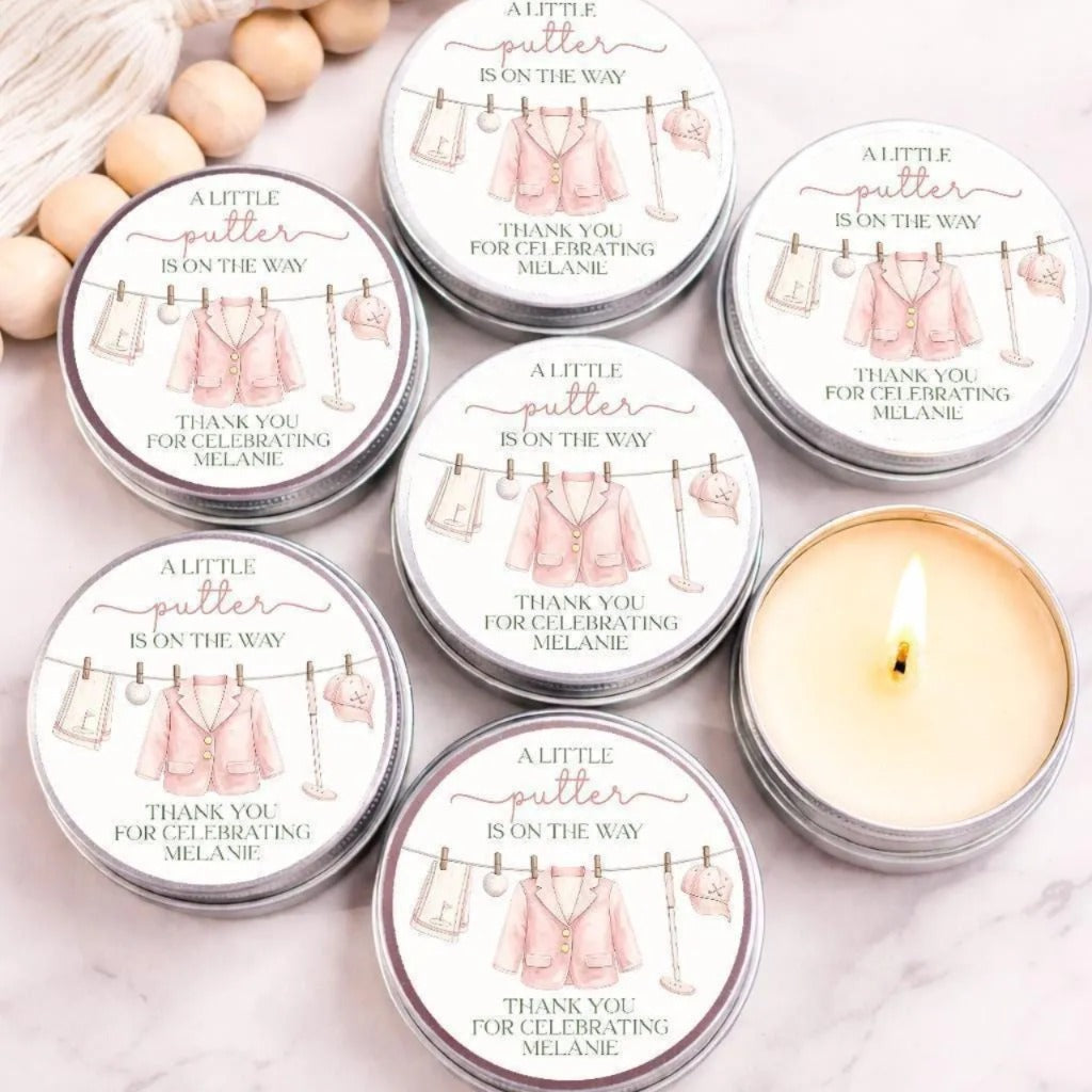 a collection of round tin candles with a pink and white color scheme, arranged on a marble surface. The candles are labeled with a message that reads "A little putter is on the way" and "Thank you for celebrating Melanie".