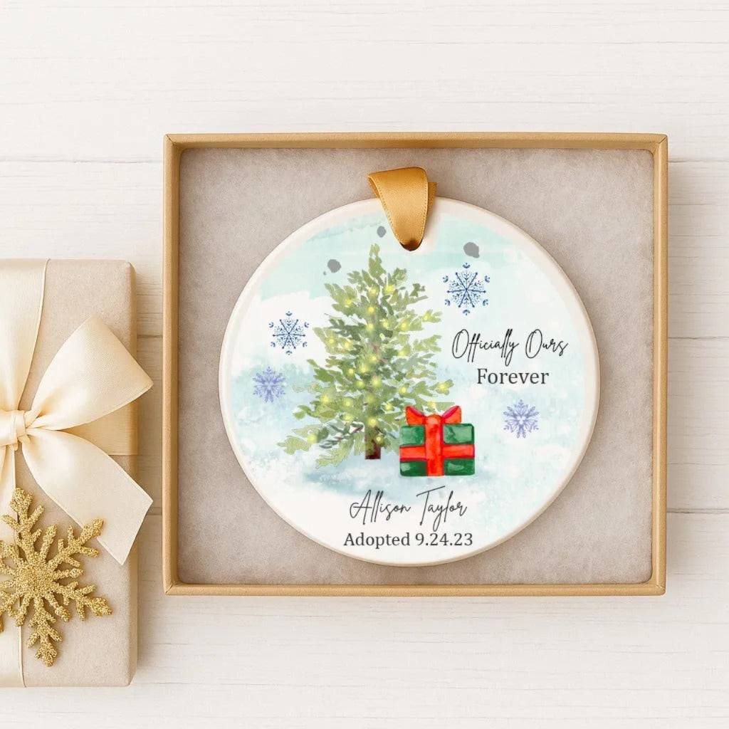 A white ceramic ornament with a Christmas tree and a red gift box, displayed in a wooden box with a gold ribbon.