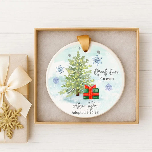 A white ceramic ornament with a Christmas tree and a red gift box, displayed in a wooden box with a gold ribbon.