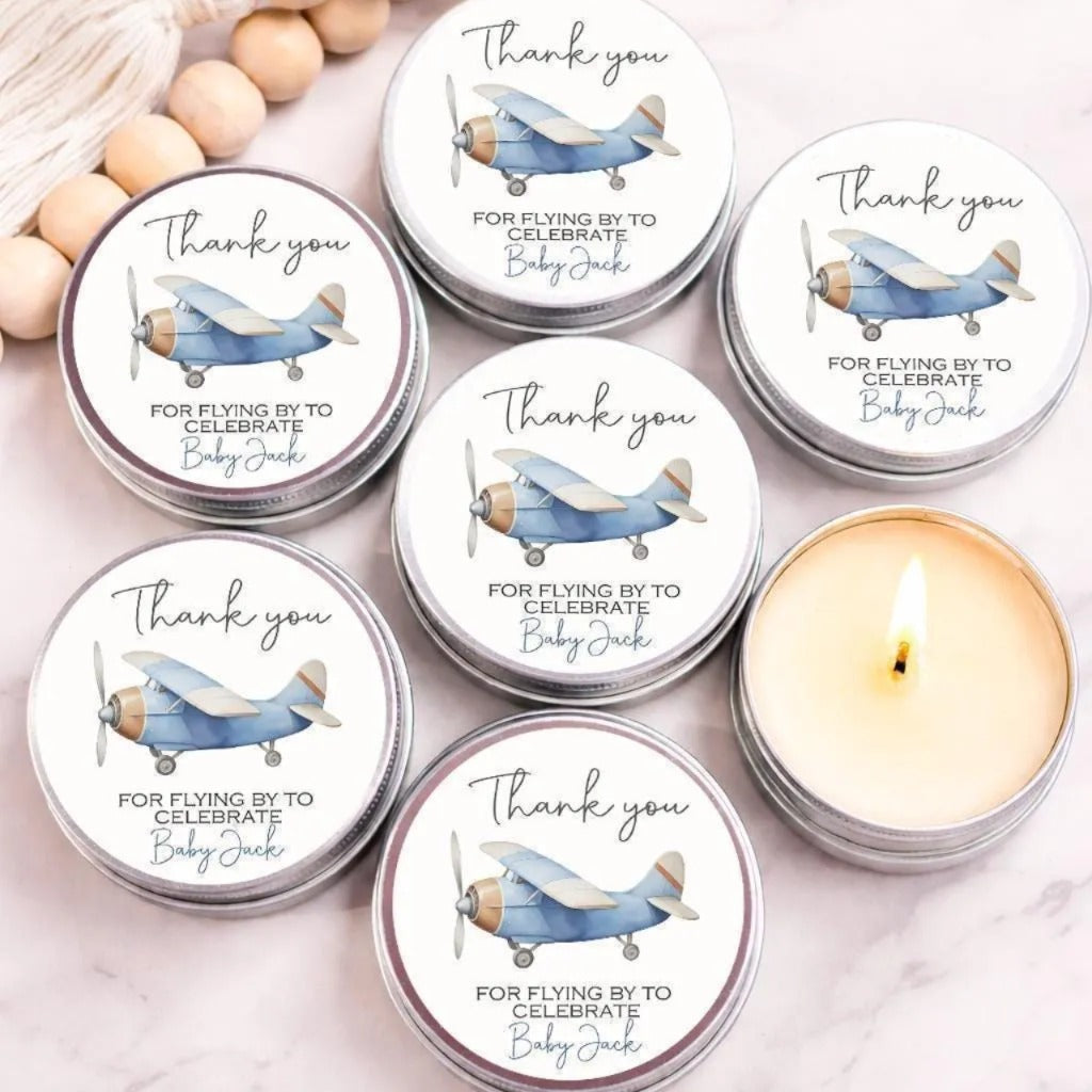 a collection of round tin candles with a blue and white airplane design, arranged on a marble surface. The candles are labeled with the phrase "Thank you for flying by to celebrate Baby Jack" and are accompanied by a candle in the foreground.