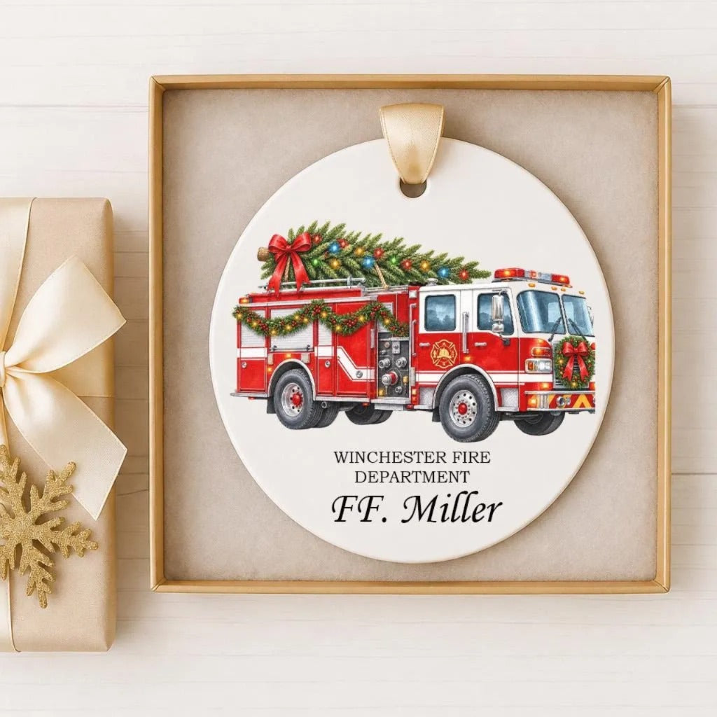 A white ceramic ornament featuring a red fire truck with Christmas decorations, displayed in a wooden frame.