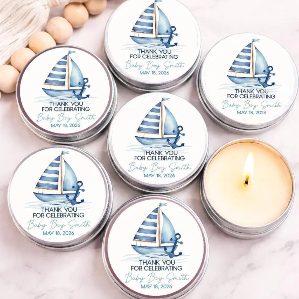 Group of personalized nautical baby shower candle favors displayed in silver tins featuring a watercolor sailboat with blue striped sails and a navy anchor. Custom labels read “Thank You for Celebrating Baby Henry” with the event date. One open candle reveals creamy wax, with decorative wooden beads and a tassel in the background.