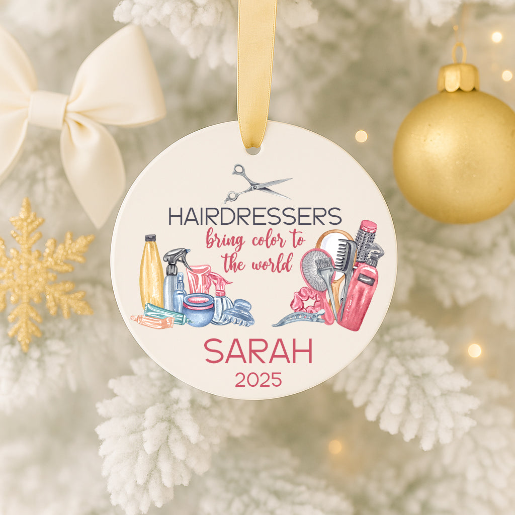A white ornament with a gold ribbon, featuring illustrations of hair styling tools and text that reads "HAIRDRESSORS bring color to the world" and "SARAH 2025".