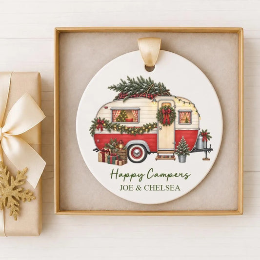 A white ceramic ornament with a red and white camper trailer decorated with Christmas lights and greenery, placed in a gold frame.