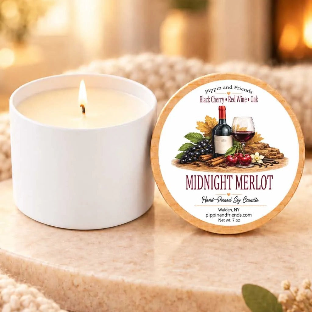 a white candle with a label that reads "Midnight Merlot" and "Pippin and Friends" next to it. The candle is placed on a wooden surface, and there is a blurred background that includes a fireplace.