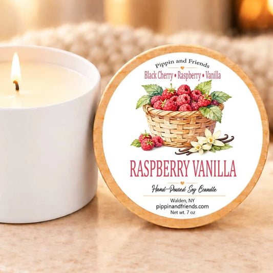 A white candle with a label that reads "RASPBERRY VANILLA" next to a lit candle on a table.