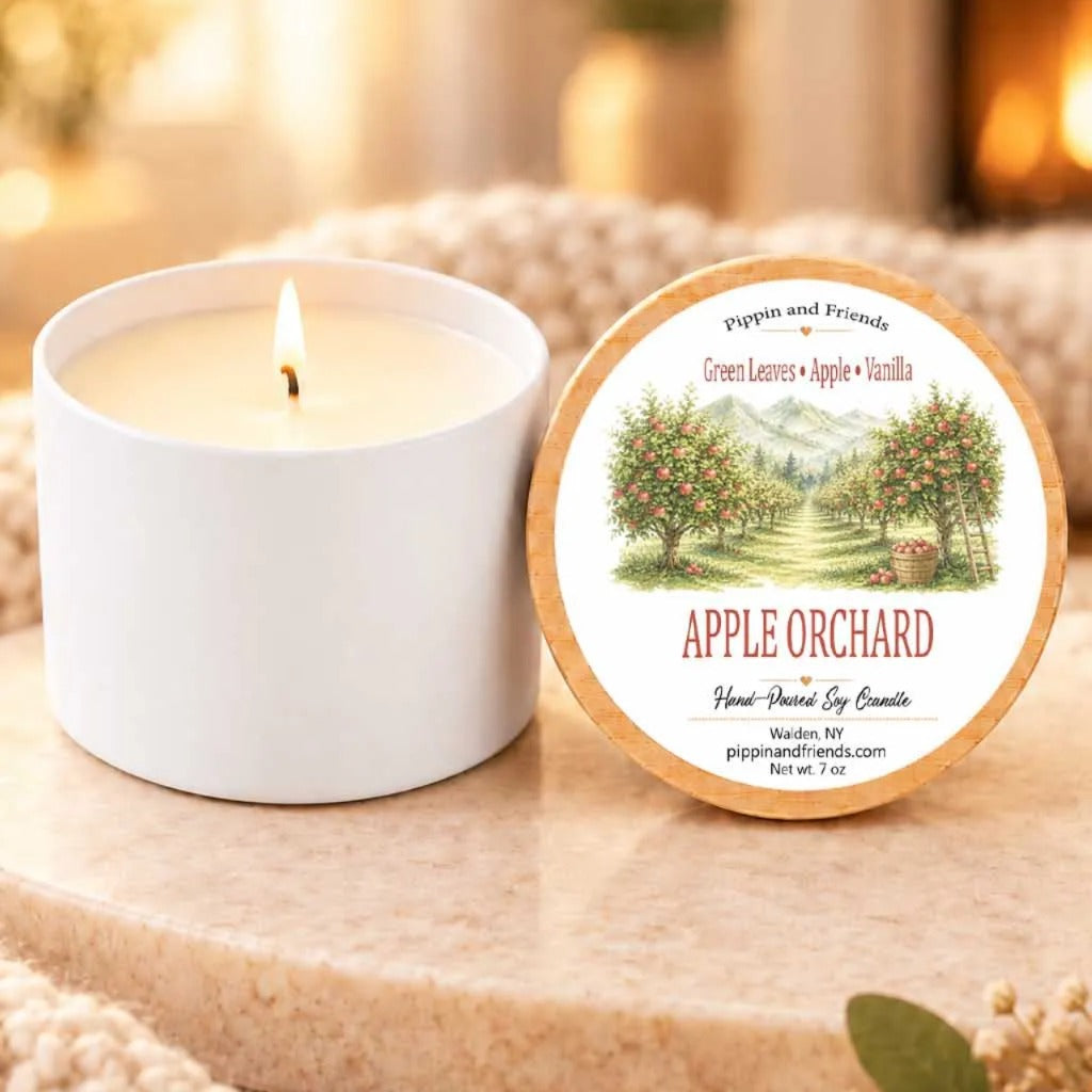 a white candle labeled "Apple Orchard" sitting on a wooden surface, with a blurred background that includes a fireplace and a window.