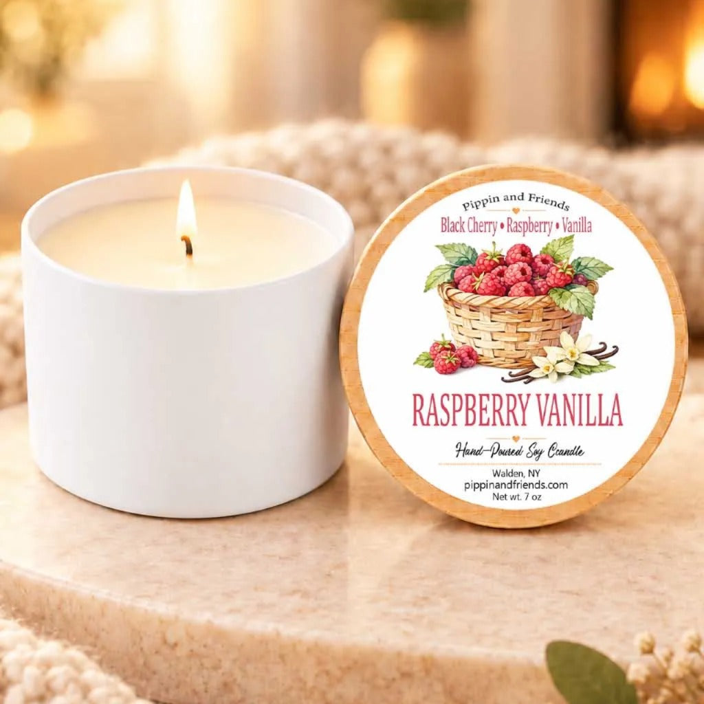 a white candle with a label that reads "RASPBERRY VANILLA" and a basket of raspberries, placed on a wooden surface next to a white candle holder.
