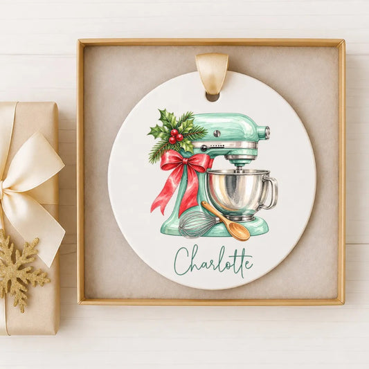 A white ceramic ornament with a turquoise mixer and a red bow, along with the name "Charlotte" written on it, is displayed in a gold frame.