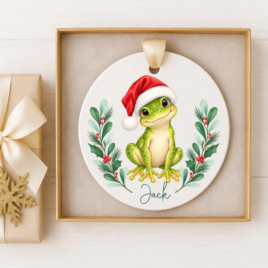 A ceramic ornament featuring a green frog wearing a red Santa hat, surrounded by holiday decorations such as holly leaves and berries.