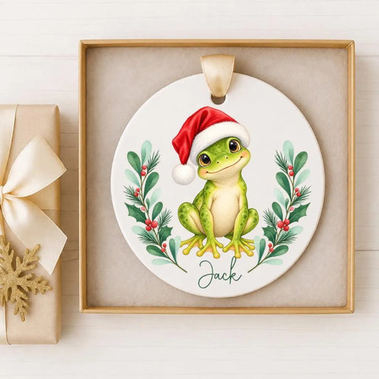 A ceramic ornament featuring a green frog wearing a red Santa hat, surrounded by holiday decorations such as holly leaves and berries.