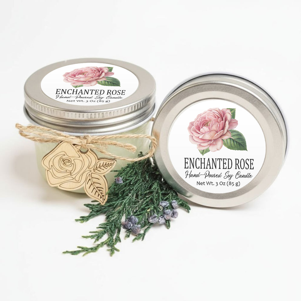 two jars of scented candles, one labeled "Enchanted Rose" and the other labeled "Enchanted Rose Hand-Dipped Soy Candle". The jars are placed next to a sprig of greenery and a small wooden tag.