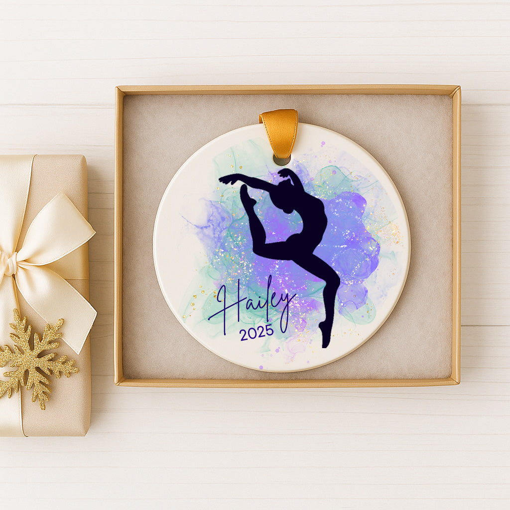 A ceramic ornament with a silhouette of a person doing a dance pose, surrounded by a watercolor splash effect. The ornament is displayed in a gold frame, and there is a gift box with a gold bow next to it.