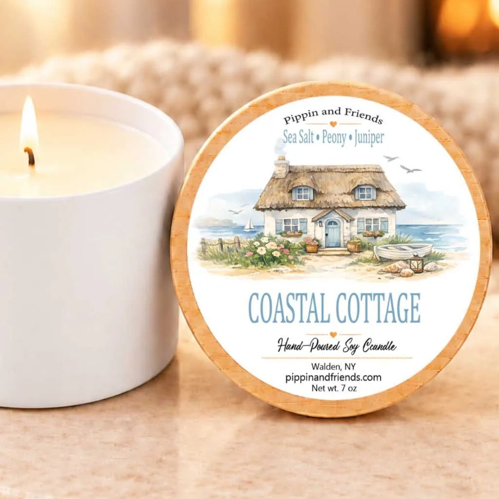 a white candle next to a round label with a coastal cottage illustration and text.