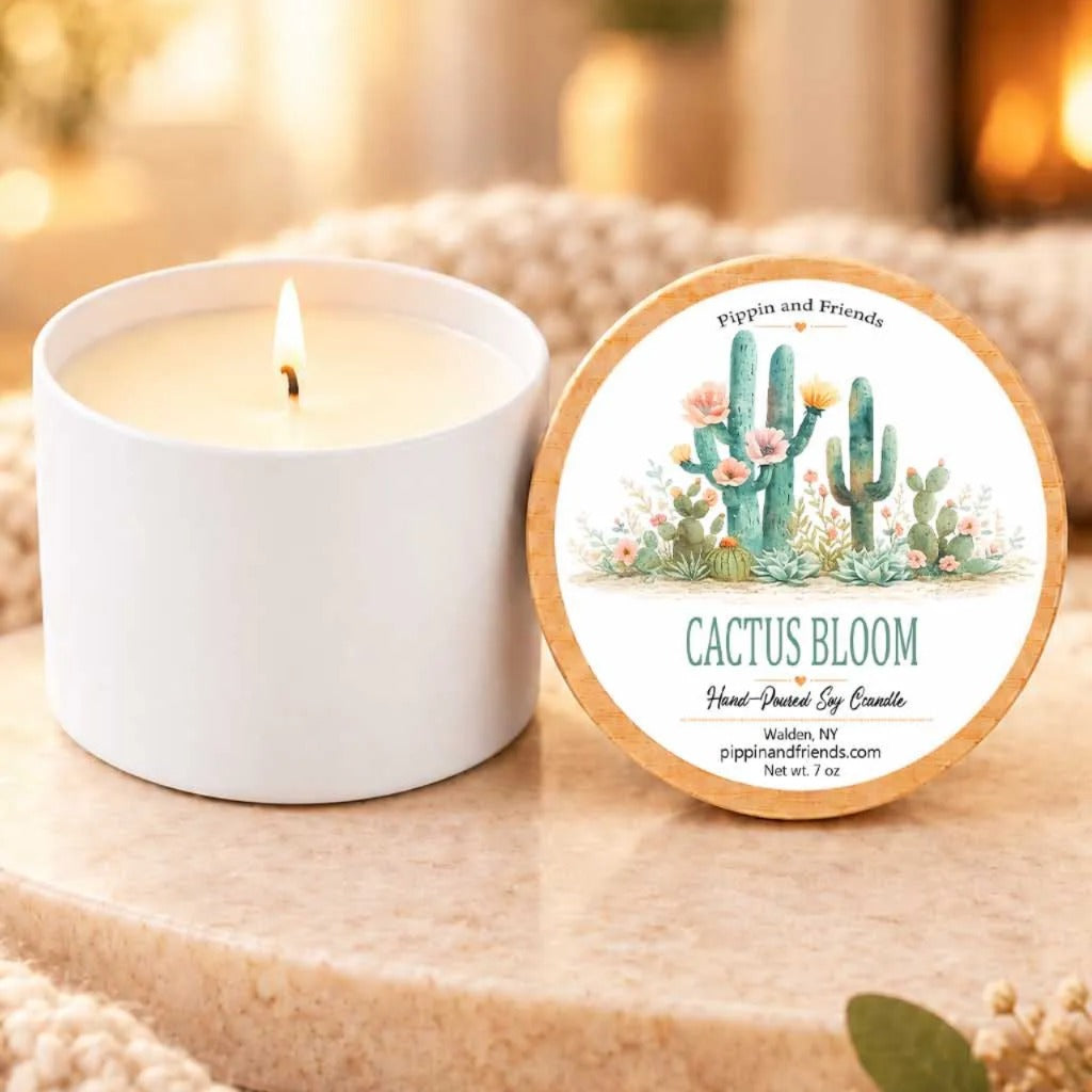 a white candle with a label that reads "Cactus Bloom" and "Pippin and Friends" on it, placed on a wooden surface next to a white candle holder.