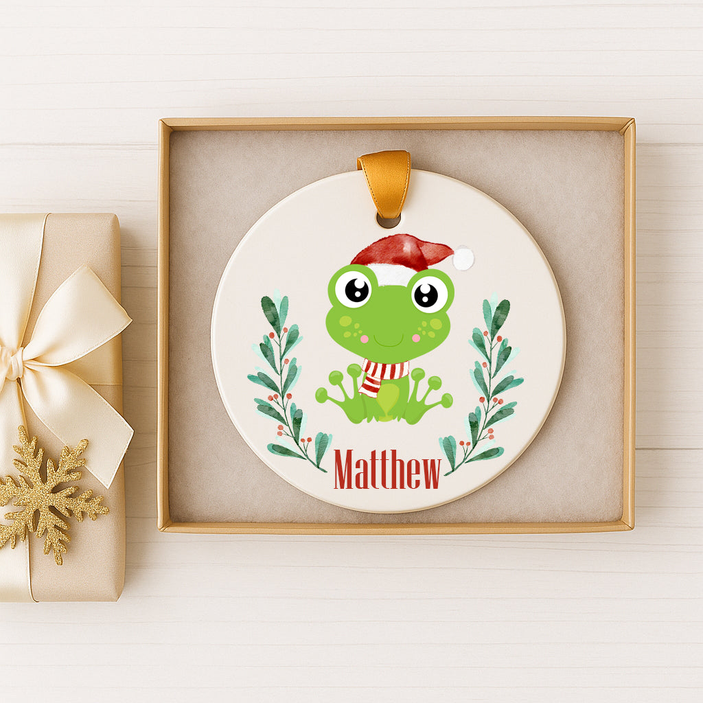 A white ceramic ornament with a green frog wearing a Santa hat, surrounded by green leaves and berries, and the name "Matthew" written below.