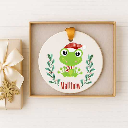 A white ceramic ornament with a green frog wearing a Santa hat, surrounded by green leaves and berries, and the name "Matthew" written below.